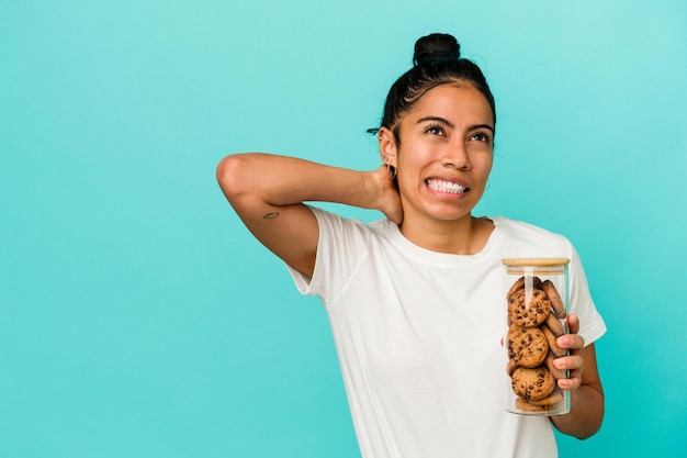 Giovane donna latina che tiene un barattolo di biscotti isolato su priorità bassa blu che tocca la parte posteriore della testa, pensando e facendo una scelta.