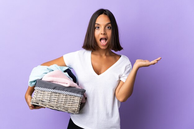 Giovane donna latina che tiene un cestino dei vestiti isolato sulla porpora con l'espressione facciale scioccata