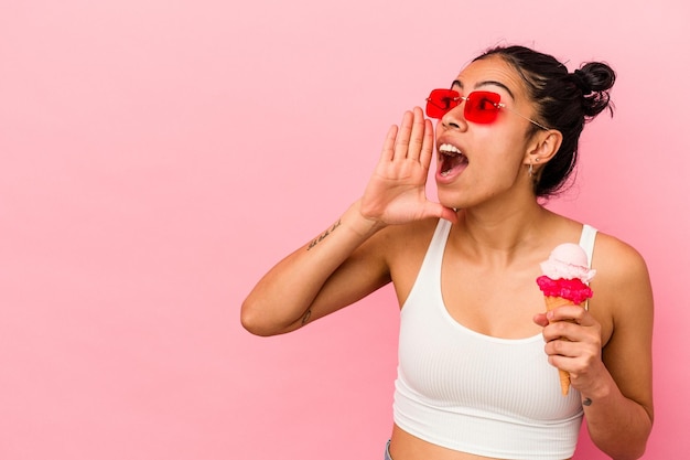 Giovane donna latina che tiene un gelato isolato su sfondo rosa gridando e tenendo il palmo vicino alla bocca aperta.