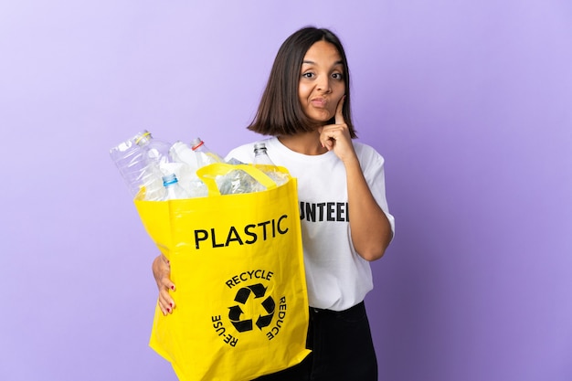 Giovane donna latina che tiene un sacchetto di riciclaggio pieno di carta da riciclare isolato sulla porpora e sul pensiero