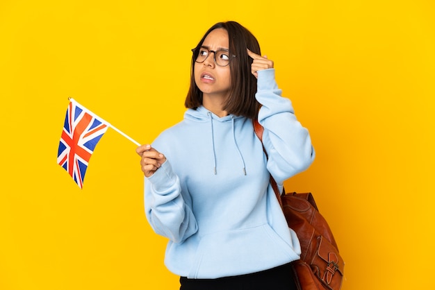 Giovane donna latina che tiene una bandiera del Regno Unito isolata su priorità bassa gialla che ha dubbi e pensiero