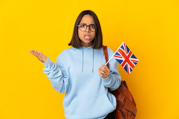 Giovane donna latina che tiene una bandiera del Regno Unito isolata sulla parete gialla che fa il gesto di dubbi