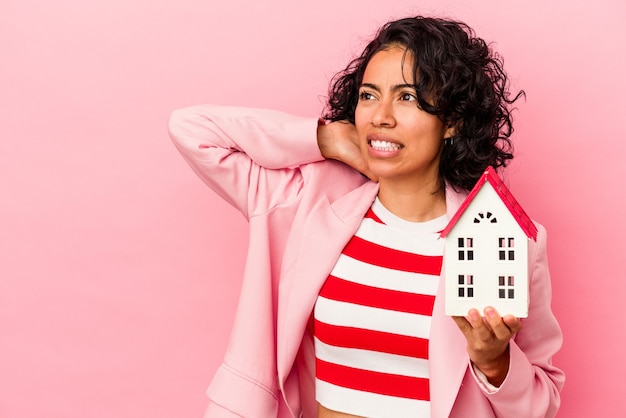 Giovane donna latina che tiene una casa giocattolo isolata su sfondo rosa toccando la parte posteriore della testa, pensando e facendo una scelta.