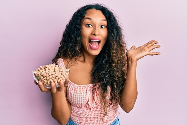 Giovane donna latina che tiene una ciotola di ceci che celebra il successo con un sorriso felice e un'espressione vincente con la mano alzata