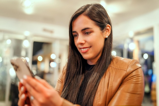 Giovane donna latina che utilizza un dispositivo mobile e sorride all'interno in un ristorante o pub portraitx9