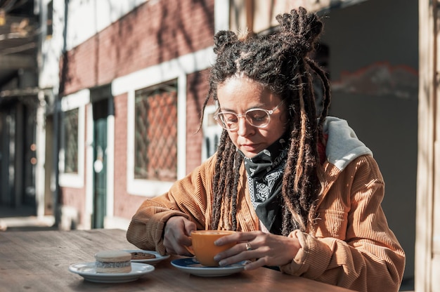 Giovane donna latina con i dreadlocks che tiene una tazza di caffè gialla fuori dalla caffetteria