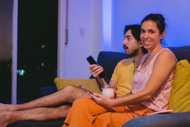 Giovane donna latina con un compagno in mano che guarda la macchina fotografica mentre condivide con un amico che guarda lo spazio della copia della TV