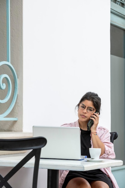 Giovane donna latina con un laptop in possesso di uno smartphone mentre si siede al caffè sul marciapiede stock foto