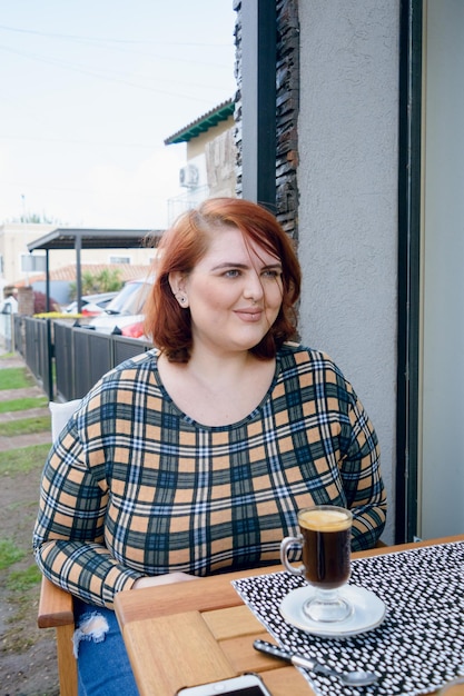 Giovane donna latina plus size seduta fuori dalla caffetteria sorridente guardando dritto davanti a sé