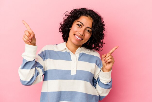 Giovane donna latina riccia isolata su sfondo rosa che punta a diversi spazi di copia, scegliendone uno, mostrando con il dito.