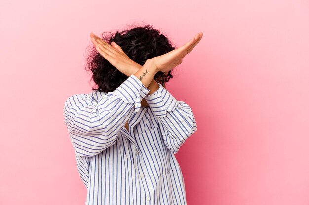 Giovane donna latina riccia isolata su sfondo rosa mantenendo due braccia incrociate, concetto di negazione.