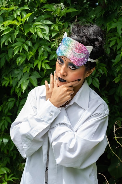 Giovane donna latina travestita con espressione seria strano trucco e maschera colorata sopra la testa su sfondo naturale