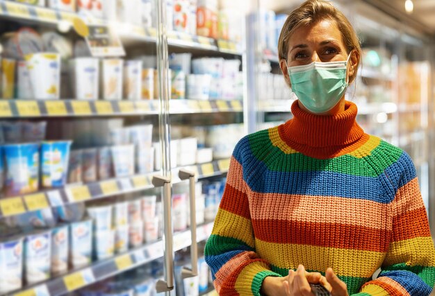 Giovane donna lesbica che indossa un maglione arcobaleno con maschera facciale che acquista generi alimentari nella sezione congelatore del supermercato durante la pandemia di Coronavirus