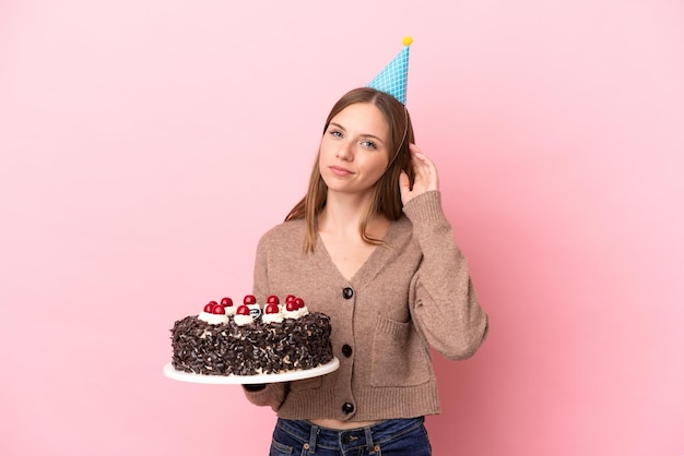 Giovane donna lituana che tiene la torta di compleanno isolata su fondo rosa che ha dubbi