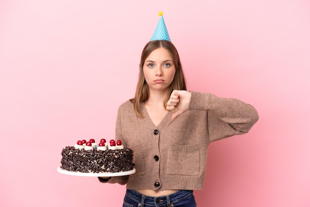 Giovane donna lituana che tiene la torta di compleanno isolata su sfondo rosa che mostra il pollice verso il basso con un'espressione negativa