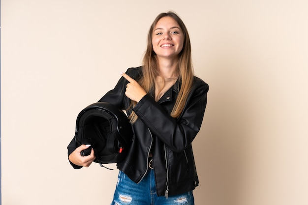 Giovane donna lituana che tiene un casco da motociclista isolato sulla parete beige che punta di lato per presentare un prodotto