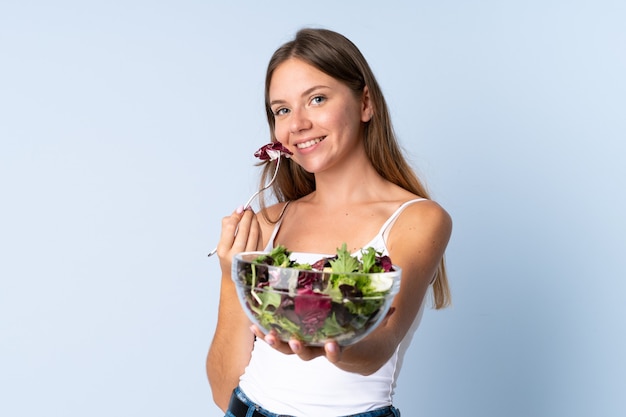 Giovane donna lituana isolata su sfondo blu, tenendo una ciotola di insalata con felice espressione