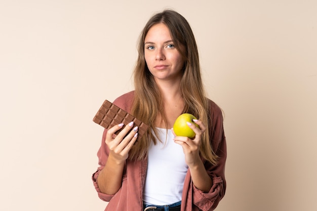 Giovane donna lituana isolata sul beige prendendo una tavoletta di cioccolato in una mano e una mela nell'altra