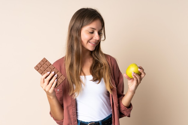 Giovane donna lituana isolata sul muro beige prendendo una tavoletta di cioccolato in una mano e una mela nell'altra
