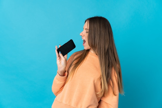 Giovane donna lituana isolata sulla parete blu utilizzando il telefono cellulare e cantando