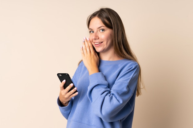 Giovane donna lituana utilizzando il telefono cellulare isolato sul muro beige sussurrando qualcosa