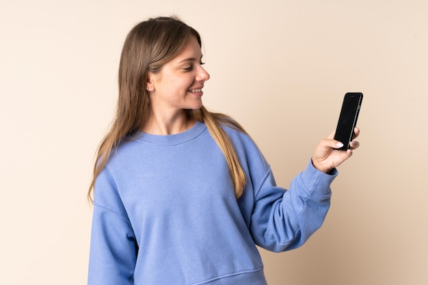 Giovane donna lituana utilizzando il telefono cellulare isolato sulla parete beige con felice espressione