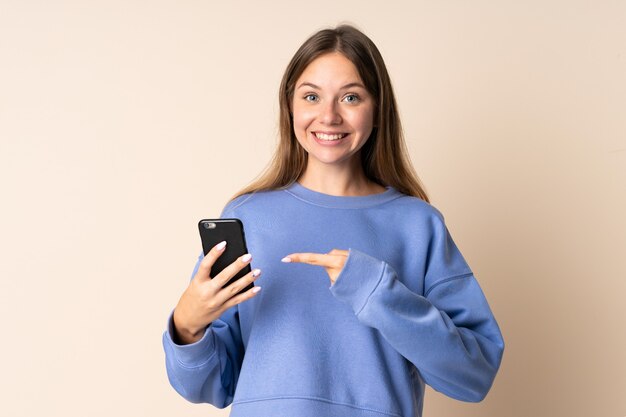 Giovane donna lituana utilizzando il telefono cellulare sul beige con espressione facciale a sorpresa