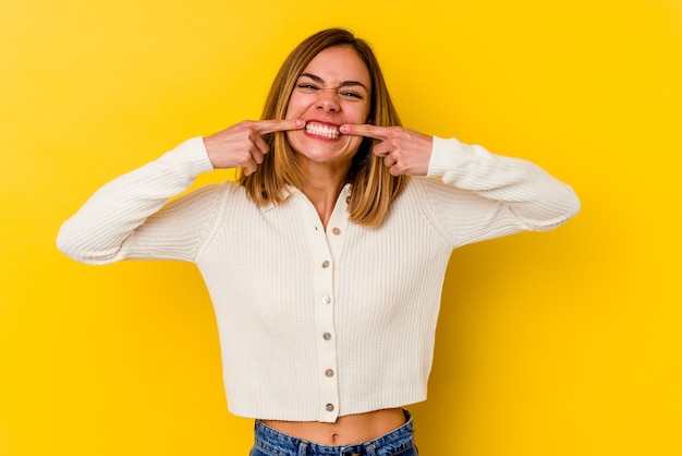 Giovane donna magra caucasica isolata sul muro giallo sorride, puntando le dita alla bocca.