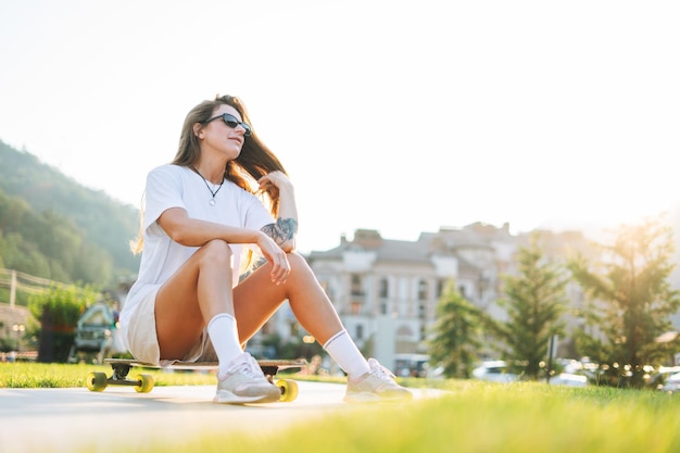 Giovane donna magra con lunghi capelli biondi in abiti sportivi leggeri che si siede con il longboard nello skatepark