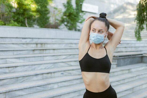 Giovane donna magra in maschera protettiva medica che fa stretching prima dell'allenamento all'aperto. Sport durante il periodo covid-19