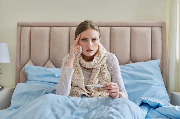 Giovane donna malata con il termometro in mano seduta a casa a letto