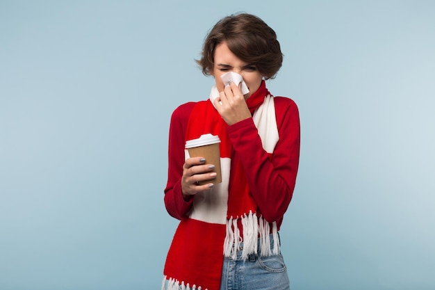 Giovane donna malsana in maglione rosso e sciarpa che tiene una tazza di caffè per andare in mano e soffia il naso con un tovagliolo su sfondo blu isolato