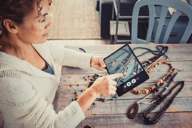 Giovane donna matura che scatta foto di collane gioielli fatti a mano a casa nel posto di lavoro dell'officina con un tablet moderno Attività per il tempo libero hobby indoor e concetto di stile di vita moderno business online