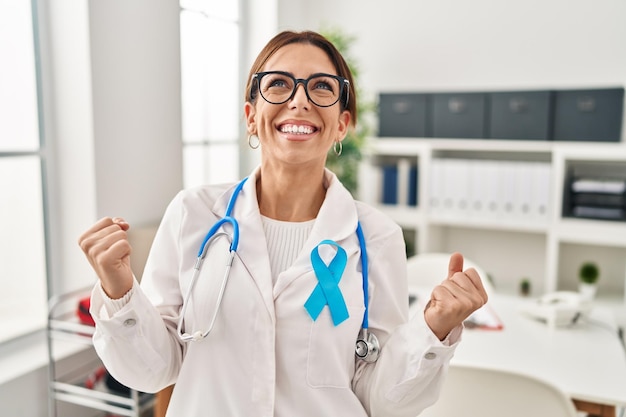 Giovane donna medico bruna che indossa uno stetoscopio presso la clinica molto felice ed eccitata facendo il gesto del vincitore con le braccia alzate, sorridendo e urlando per il successo. concetto di celebrazione.