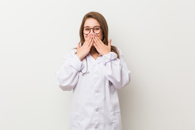 Giovane donna medico contro un muro bianco ridendo di qualcosa, coprendo la bocca con le mani.