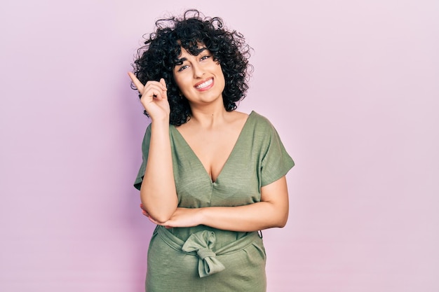 Giovane donna mediorientale che indossa abiti casual con un grande sorriso sul viso che punta con la mano e il dito di lato guardando la telecamera