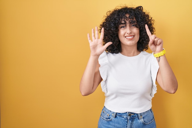 Giovane donna mediorientale in piedi su sfondo giallo che mostra e punta verso l'alto con le dita numero sei mentre sorride fiduciosa e felice