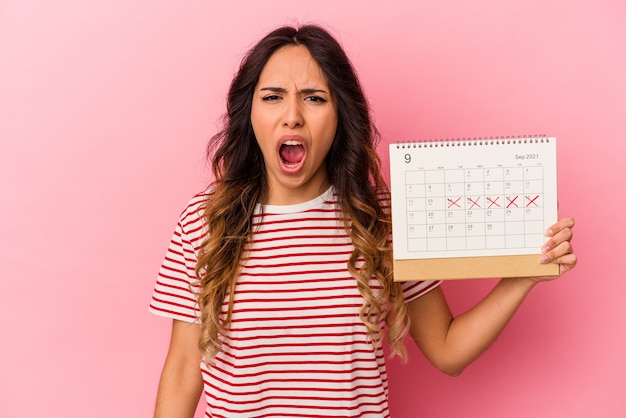 Giovane donna messicana che tiene un calendario isolato sulla parete rosa che grida molto arrabbiato e aggressivo.