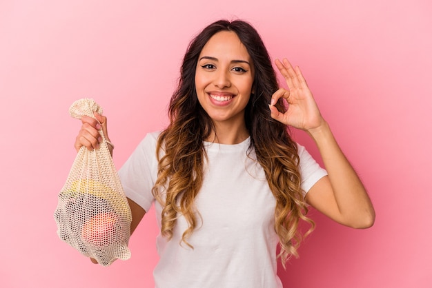 Giovane donna messicana che tiene un sacchetto di frutta isolato sulla parete rosa allegro e fiducioso che mostra gesto giusto.