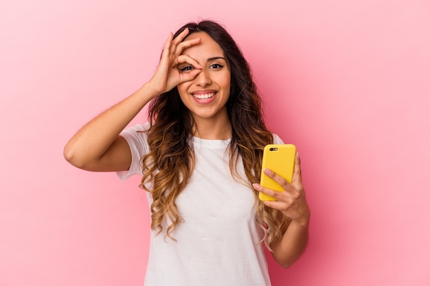 Giovane donna messicana che tiene un telefono cellulare isolato sul rosa eccitato mantenendo il gesto giusto sull'occhio.