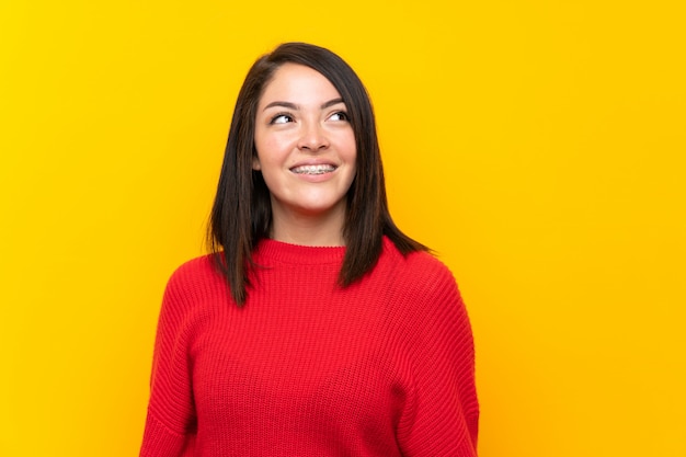 Giovane donna messicana con maglione rosso oltre muro giallo ridendo e alzando lo sguardo