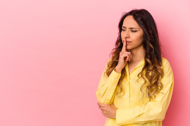 Giovane donna messicana isolata sulla parete rosa che guarda lateralmente con espressione dubbiosa e scettica.