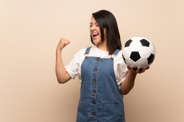 Giovane donna messicana sopra la parete isolata che tiene un pallone da calcio