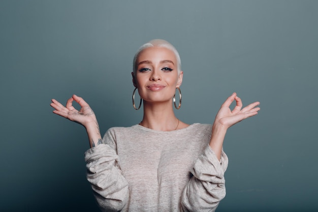 Giovane donna millenaria con i capelli biondi corti ritratto seduto e meditare con il segno ok mudra su sfondo grigio blu