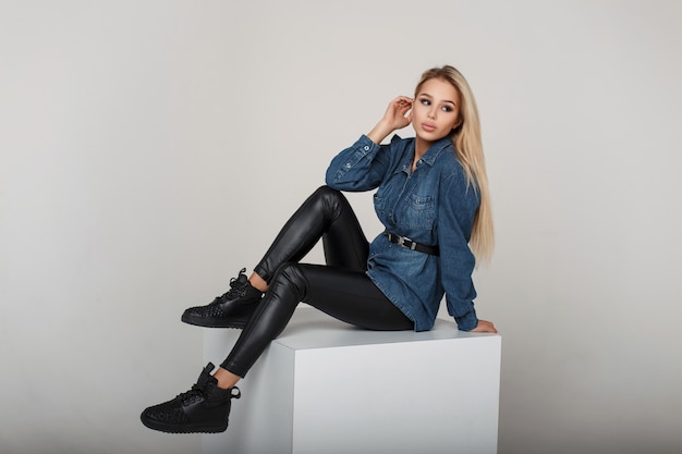 Giovane donna modello americano in camicia di jeans e pantaloni neri con scarpe da ginnastica alla moda nere si siede in studio
