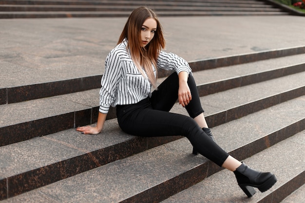 Giovane donna moderna in jeans alla moda in una camicia a righe alla moda in scarpe di pelle nera in posa su una scala d'epoca in città in una calda giornata di primavera all'aperto