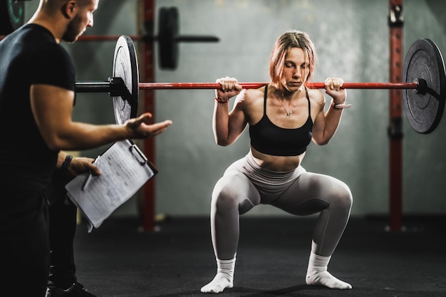 Giovane donna muscolare che lavora con il personal trainer in palestra. Sta facendo esercizi di squat con il bilanciere.