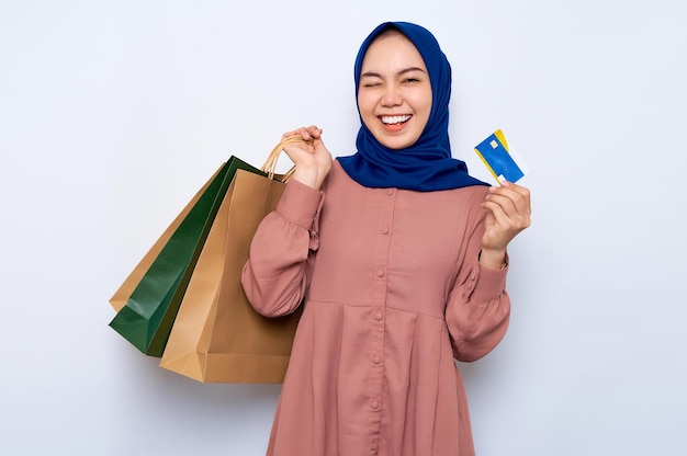 Giovane donna musulmana asiatica allegra in camicia rosa che tiene borse per pacchetti con acquisti dopo lo shopping e carta di credito isolata su sfondo bianco Concetto di stile di vita delle persone