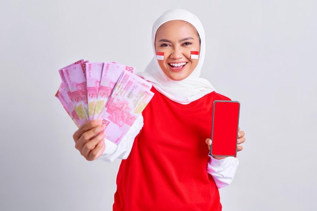 Giovane donna musulmana asiatica allegra in maglietta bianca rossa che mostra telefono cellulare e denaro contante in banconote in rupia indonesiana isolate su sfondo bianco Festa dell'indipendenza indonesiana il 17 agosto concetto