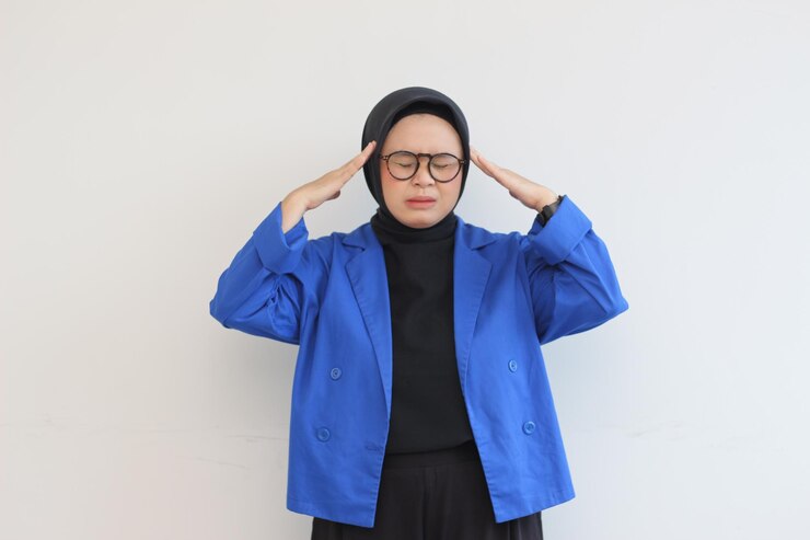 giovane donna musulmana asiatica che indossa occhiali e blazer blu con espressione vertiginosa che tiene la testa